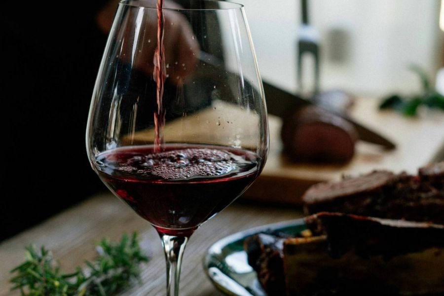 Red wine being poured into a glass, with someone slicing meat in the background on a wooden table.