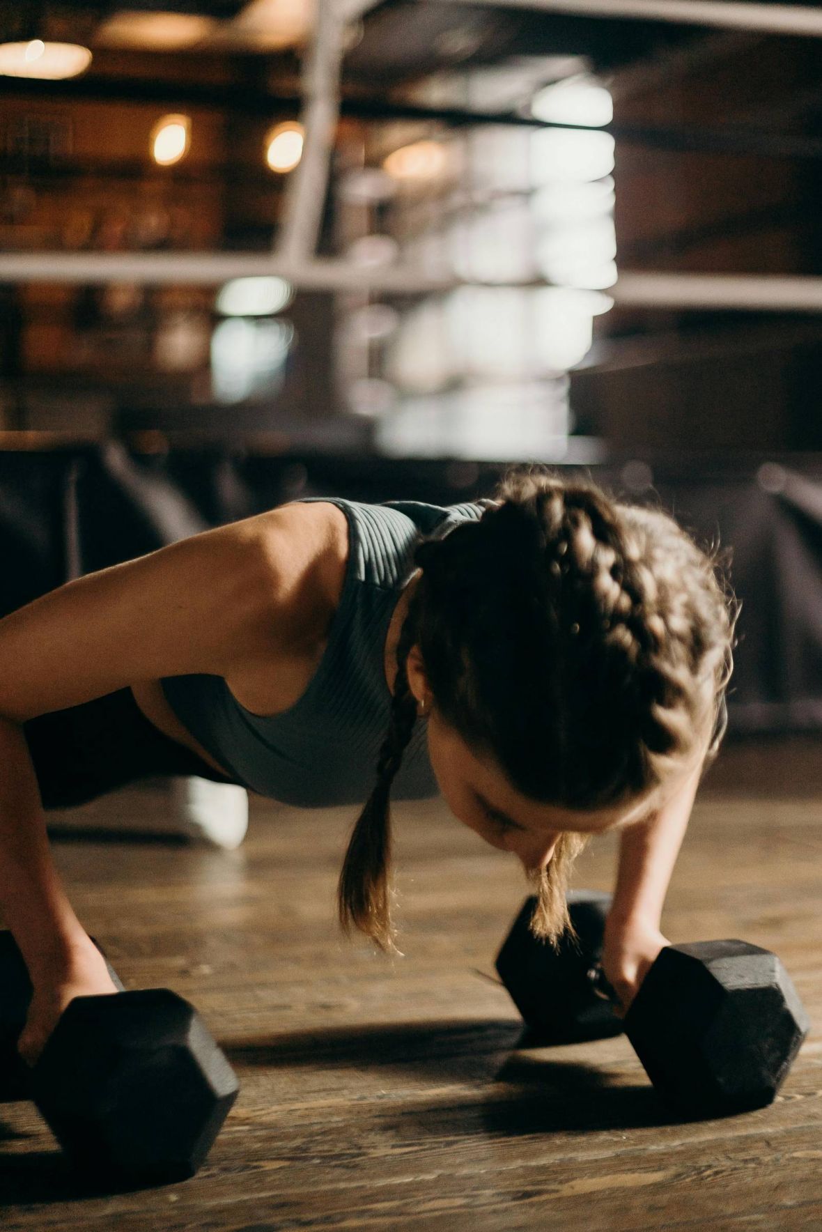 Woman with braided hair doing push-ups on dumbbells in a gym with blurred boxing ring in background.