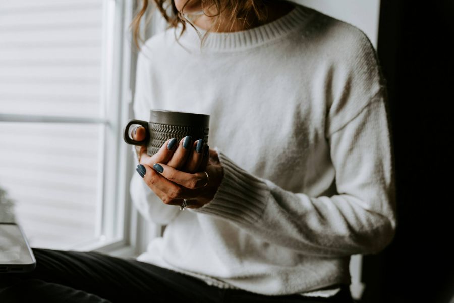 Woman in a white sweater holding a mug with both hands, sitting by a window.