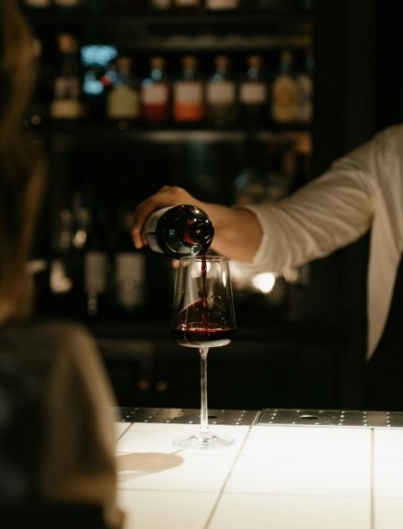 A person pours red wine into a glass at a dimly lit bar with bottles in the background.