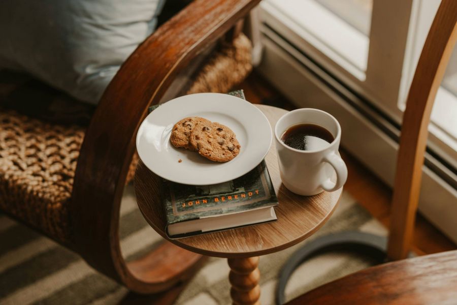 A plate with two cookies, a cup of coffee, and a book on a small wooden table next to a rocking chair.